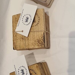 May include: Three sets of square wooden coasters, each tied with twine and featuring a tag. The tags display a crown logo and the words "CROWN FABRICATION". The coasters are made of light brown wood.