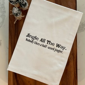 May include: White kitchen towel with the text "Jingle All The Way. Nobody likes a half-assed jingler." displayed in black font. The towel is placed on a wooden cutting board, with three small, snow-covered decorative pine trees in the upper left corner.
