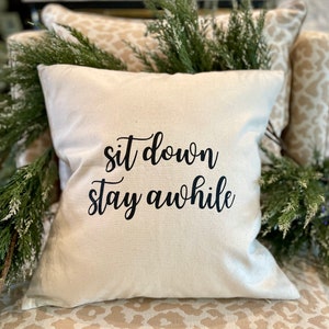 May include: A white throw pillow with the text "sit down stay awhile" in black cursive lettering.