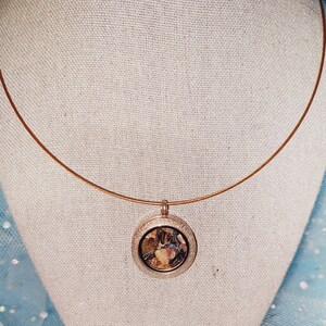 May include: A copper-colored necklace with a round pendant. The pendant has a silver-colored frame and contains a mix of brown and beige elements. The necklace is displayed on a white mannequin head against a light blue background.