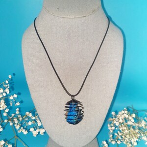 May include: A blue and brown stone encased in a black wire cage pendant on a black cord necklace. The necklace is displayed on a gray mannequin bust.