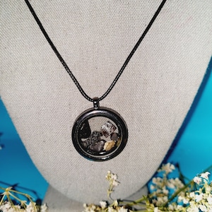 May include: A black necklace with a round pendant containing small black, brown, and gold stones. The necklace is displayed on a gray mannequin against a blue background.