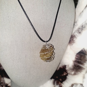 May include: A pendant necklace featuring a raw, light-yellow crystal wrapped in silver-colored wire. The crystal hangs from a black cord necklace, displayed against a neutral background and a fluffy, brown and white patterned surface.
