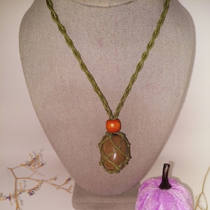 May include: A handmade necklace with a brown stone pendant encased in a green woven net. The necklace has a green cord and an orange bead. Two purple fabric pumpkins with brown stems are in the foreground.