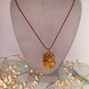 May include: A gold wire wrapped pendant necklace featuring a large, natural, orange-brown crystal. The necklace is on a brown cord and is displayed on a gray mannequin bust.