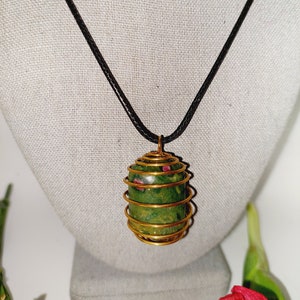 May include: A green and red stone pendant necklace. The stone is encased in a gold-colored wire cage and hangs from a black cord. The pendant is displayed on a gray mannequin bust.