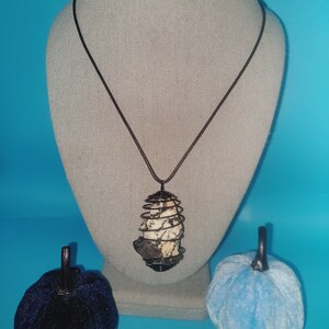 May include: A brown leather cord necklace with a caged crystal pendant. The crystal is white and gray with black accents. The necklace is displayed on a white mannequin.