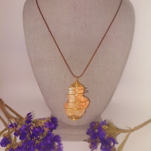 May include: A gold wire wrapped pendant necklace featuring a light orange crystal. The necklace is on a brown cord and is displayed on a fabric mannequin.