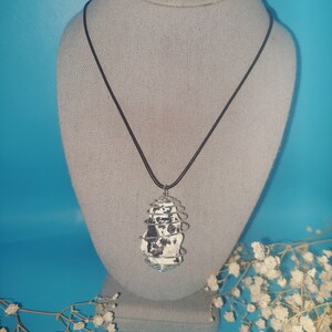 May include: A black and white stone pendant encased in a silver wire cage, hanging from a black cord necklace. The necklace is displayed on a gray fabric mannequin.