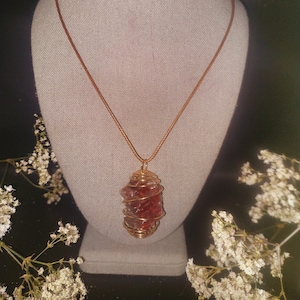 May include: A red and clear crystal pendant wrapped in gold wire, hanging from a brown cord necklace.