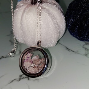 May include: A silver locket necklace with a clear glass insert containing small pink and purple crystals. The necklace is on a white fuzzy surface.