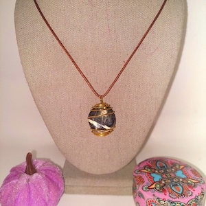 May include: A handcrafted necklace featuring a wire-wrapped stone pendant with a brown cord. The pendant is a sphere of dark stone with white veining, encased in gold-colored wire. A small, pink, velvet pumpkin and a decorative pink box are also in the image.