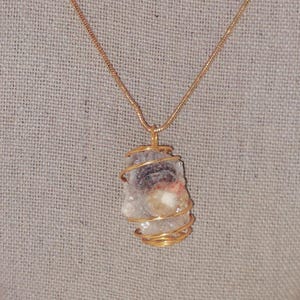 Puede incluir: Un collar con cadena dorada y colgante. El colgante es una piedra en bruto de forma irregular, envuelta en alambre dorado. La piedra tiene una mezcla de colores, incluyendo blanco, gris y toques de rosa y marr&oacute;n.