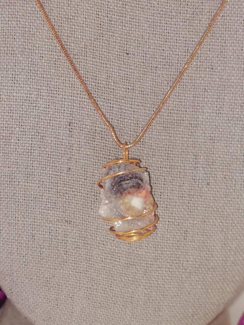 Puede incluir: Un collar con cadena dorada y colgante. El colgante es una piedra en bruto de forma irregular, envuelta en alambre dorado. La piedra tiene una mezcla de colores, incluyendo blanco, gris y toques de rosa y marr&oacute;n.