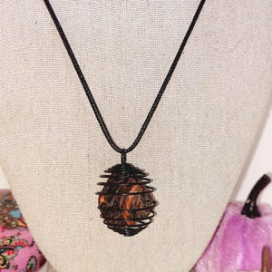 May include: A pendant necklace featuring a wire cage design in black, encasing a stone with orange and brown hues. The necklace has a black cord and is displayed on a neutral-colored surface.