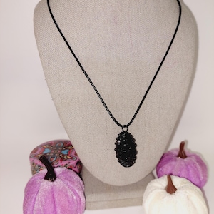 May include: A black pendant necklace with an oval-shaped, textured pendant. The necklace is displayed on a mannequin bust. Decorative purple and white velvet pumpkins are in the foreground.