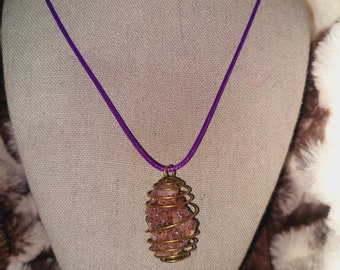 May include: A purple cord necklace with a pendant featuring a cluster of pink crystals encased in a gold-colored wire cage. The necklace is displayed on a mannequin with a neutral-colored fabric.
