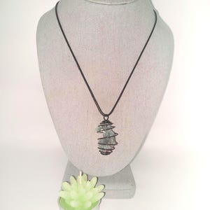 May include: A pendant necklace with a green stone wrapped in dark wire, displayed on a gray mannequin. The necklace hangs from a black cord. A small, light green candle in a silver holder sits below the mannequin.