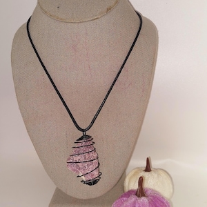 May include: A pink stone pendant necklace with a black wire wrap and cord. The pendant is displayed on a mannequin bust. Two decorative pumpkins, one white and one purple, are in the foreground.