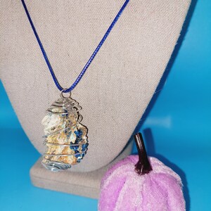 May include: A silver wire wrapped pendant necklace with a blue and white crystal inside. The necklace is on a blue background with a purple velvet pumpkin.