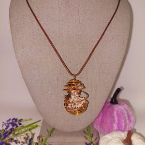 May include: A handcrafted necklace featuring a wire-wrapped stone pendant. The pendant is a light brown color with a spiral wire design. The necklace has a brown cord and is displayed on a mannequin.