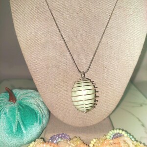 May include: A silver-toned necklace with a light green, oval-shaped pendant encased in a cage-like design. The pendant hangs from a delicate chain. A teal velvet pumpkin and a collection of colorful beaded jewelry are in the background.
