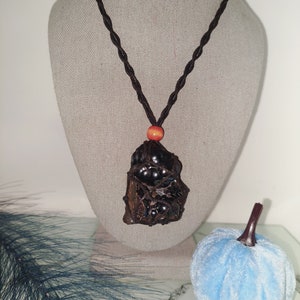 May include: A black obsidian pendant with a brown cord necklace. The pendant is a rough, irregular shape with a cluster of small black beads in the center. The cord has a small orange wooden bead near the pendant.