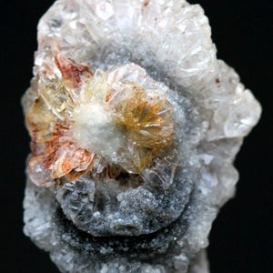 Puede incluir: Primer plano de un esp&eacute;cimen mineral con un grupo central de cristales naranjas y blancos, rodeado por una base gris texturizada. Los cristales tienen un patr&oacute;n radial, con un fondo oscuro.