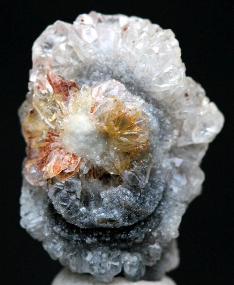 Puede incluir: Primer plano de un esp&eacute;cimen mineral con un grupo central de cristales naranjas y blancos, rodeado por una base gris texturizada. Los cristales tienen un patr&oacute;n radial, con un fondo oscuro.