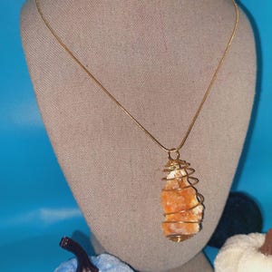 May include: A gold chain necklace with a pendant featuring a raw orange crystal encased in a gold wire spiral.