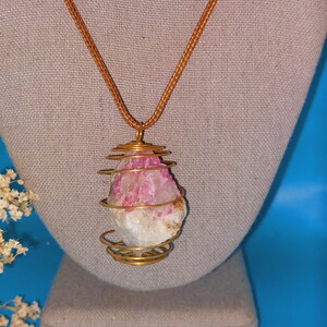 May include: A gold wire wrapped pendant necklace featuring a pink and white crystal. The necklace has a brown cord.