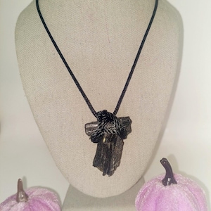 May include: A dark brown, textured, rectangular-shaped pendant suspended from a thin black cord. The pendant is secured to the cord with a black braided wrap. The piece features a rough, natural surface with vertical striations and a deep brown hue.