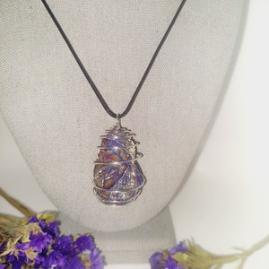 May include: A pendant necklace featuring a wire-wrapped stone with iridescent colors of blue, purple, and gold. The stone is suspended from a black cord. The necklace is displayed on a mannequin with purple flowers in the foreground.