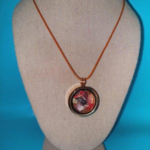 May include: A brown leather cord necklace with a round, gold-toned locket pendant. The locket contains a variety of small, colorful stones.