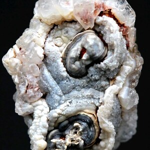 Puede incluir: Primer plano de una formaci&oacute;n mineral natural con una estructura compleja y estratificada. El mineral presenta una mezcla de tonos blancos, grises y marr&oacute;n rojizo, con estructuras cristalinas y patrones arremolinados. La textura general es rugosa y org&aacute;nica, con fondo oscuro.