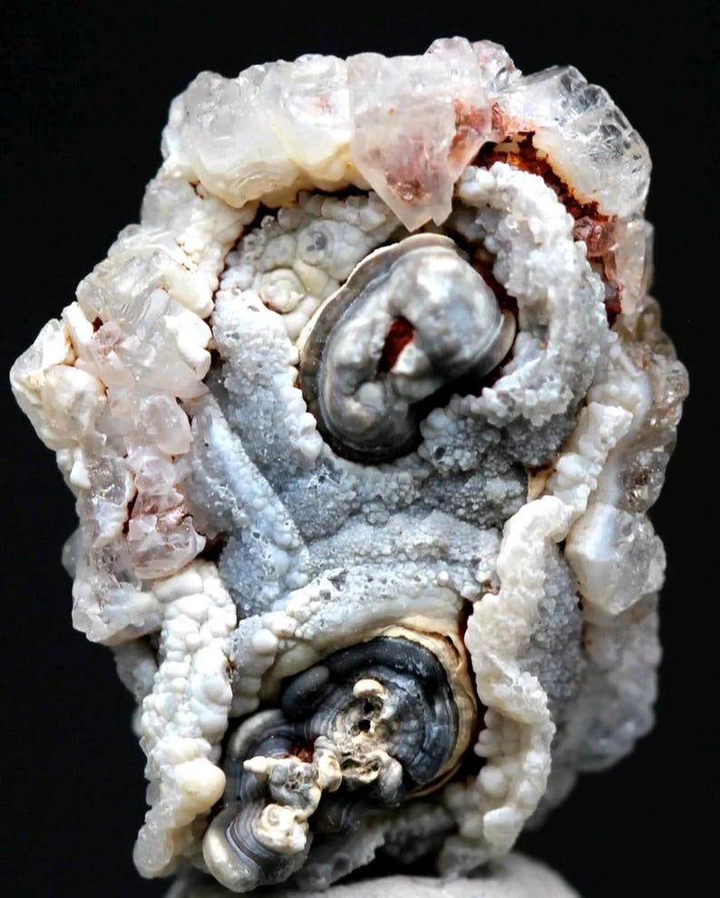 Puede incluir: Primer plano de una formaci&oacute;n mineral natural con una estructura compleja y estratificada. El mineral presenta una mezcla de tonos blancos, grises y marr&oacute;n rojizo, con estructuras cristalinas y patrones arremolinados. La textura general es rugosa y org&aacute;nica, con fondo oscuro.