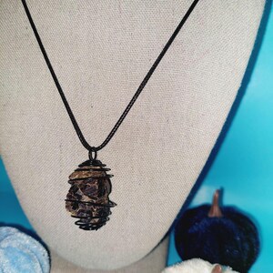 May include: A black cord necklace with a brown and black stone pendant wrapped in wire. The stone is a rough, natural shape.