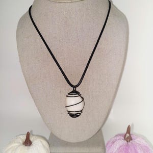 May include: A pendant necklace featuring a white, oval-shaped stone wrapped in black wire. The stone hangs from a black cord. The necklace is displayed on a mannequin. Two decorative pumpkins, one white and one lavender, are in the foreground.