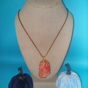 May include: A necklace featuring a raw, textured red stone pendant encased in a gold-toned wire cage, suspended from a thin brown cord. The pendant is displayed on a neutral-colored bust, flanked by a dark blue velvet pumpkin and a light blue velvet pumpkin.
