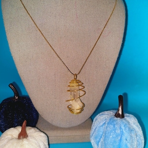 May include: A gold chain necklace with a clear quartz crystal pendant encased in a gold wire spiral. The necklace is displayed on a mannequin with a light brown fabric covering.