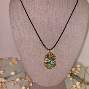 May include: A gold wire wrapped pendant necklace with a green and blue stone inside. The necklace is on a black cord and is displayed on a fabric mannequin.