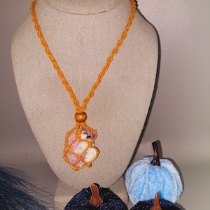 May include: A crocheted necklace with a pink and white stone pendant. The necklace is orange and the stone is in a crocheted net. The necklace is displayed on a mannequin with two blue velvet pumpkins in the foreground.