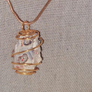 Puede incluir: Un collar con colgante artesanal con una piedra natural envuelta en alambre dorado. La piedra tiene una base blanca con patrones rojos y grises. La cadena del collar es dorada, creando un accesorio elegante y coordinado.