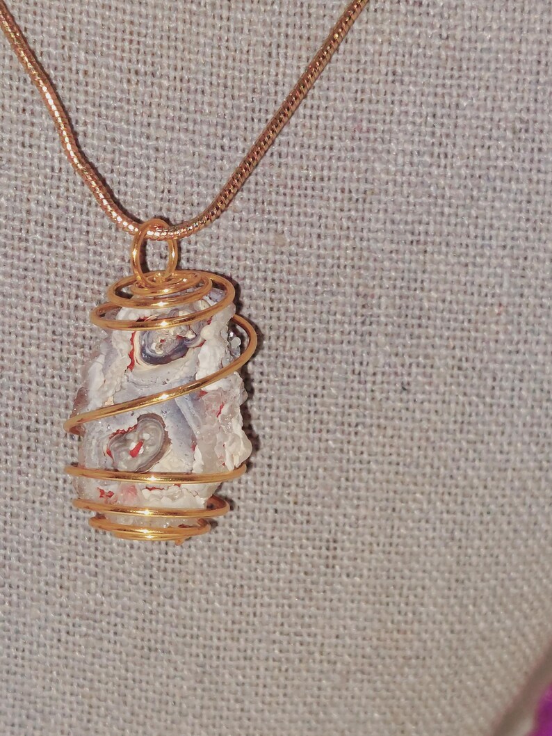 Puede incluir: Un collar con colgante artesanal con una piedra natural envuelta en alambre dorado. La piedra tiene una base blanca con patrones rojos y grises. La cadena del collar es dorada, creando un accesorio elegante y coordinado.