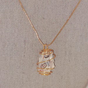 Puede incluir: Un collar con colgante envuelto en alambre dorado. El colgante presenta una piedra de color claro con marcas rojas y marrones. La piedra est&aacute; encerrada en una espiral de alambre dorado y cuelga de una delicada cadena dorada.