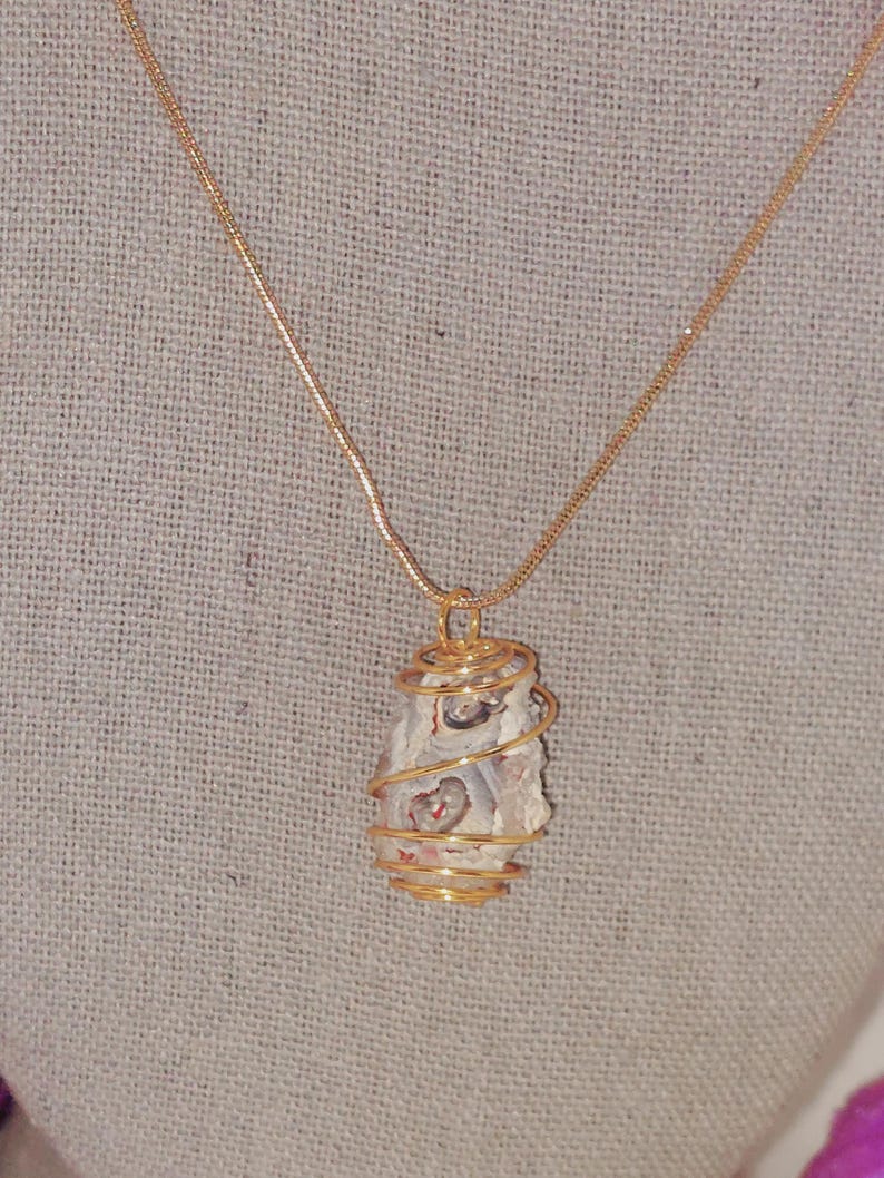 Puede incluir: Un collar con colgante envuelto en alambre dorado. El colgante presenta una piedra de color claro con marcas rojas y marrones. La piedra est&aacute; encerrada en una espiral de alambre dorado y cuelga de una delicada cadena dorada.