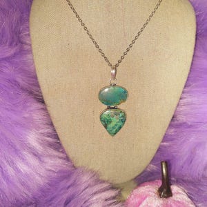 May include: A silver-toned necklace with a pendant featuring two turquoise-colored stones. The top stone is oval, and the bottom is heart-shaped. The necklace is displayed on a beige mannequin head against a purple background.
