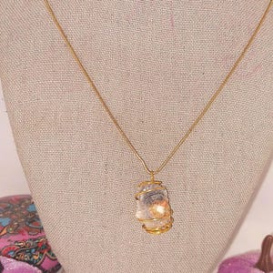 Puede incluir: Un collar con colgante envuelto en alambre dorado. El colgante es una piedra de color claro, de forma irregular, con un envoltorio de alambre dorado. El collar se exhibe en un maniqu&iacute; con una tela de color neutro.
