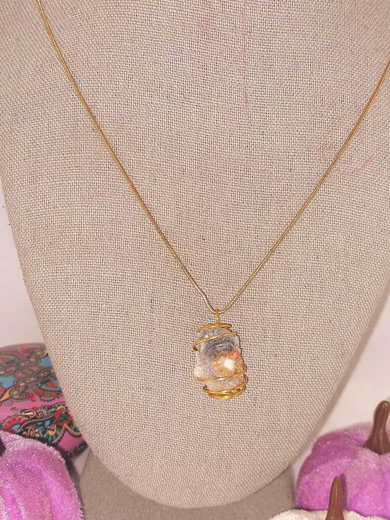 Puede incluir: Un collar con colgante envuelto en alambre dorado. El colgante es una piedra de color claro, de forma irregular, con un envoltorio de alambre dorado. El collar se exhibe en un maniqu&iacute; con una tela de color neutro.