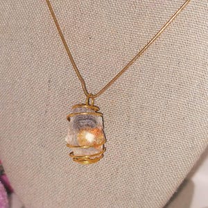 Puede incluir: Un collar con colgante envuelto en alambre dorado. El colgante presenta una piedra de color claro con inclusiones naranjas y blancas. La cadena del collar es dorada y el fondo es de tela neutra.